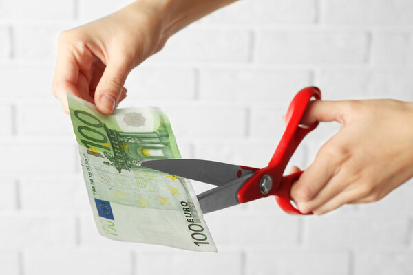 Руки с ножницами режут деньги на фоне стены из белого кирпича
