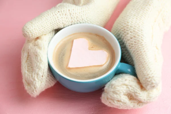 Mãos femininas em mitenes quentes segurando xícara de cappuccino quente com marshmallow coração no fundo rosa, close up — Fotografia de Stock