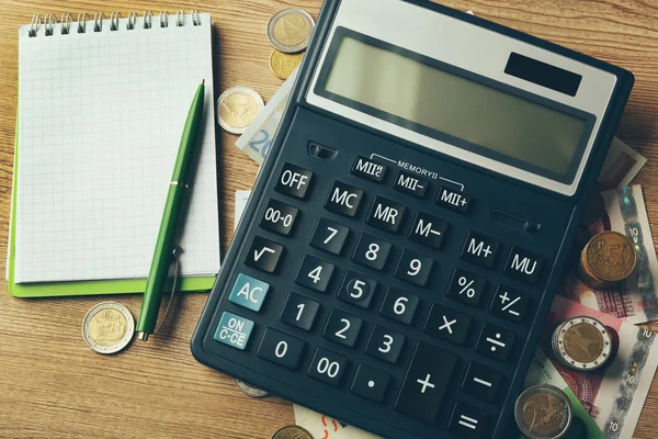 Koncept peněz. Černá kalkulačka s bankovkami a mincemi na dřevěném stole, zblizka — Stock fotografie