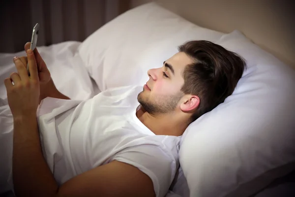 Young man lying on bed with mobile phone in bedroom — Stock Photo, Image