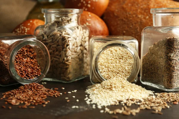 Composition of seeds in banks, bread, buns and oil on wooden table background, closeup — Stock Photo, Image