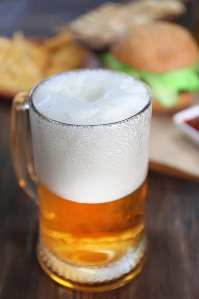 Glass mug of light beer with snacks on dark wooden table, close up — Stock Photo, Image