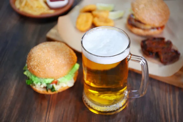 Taza de vidrio de cerveza ligera con hamburguesas en mesa de madera oscura, de cerca — Foto de Stock