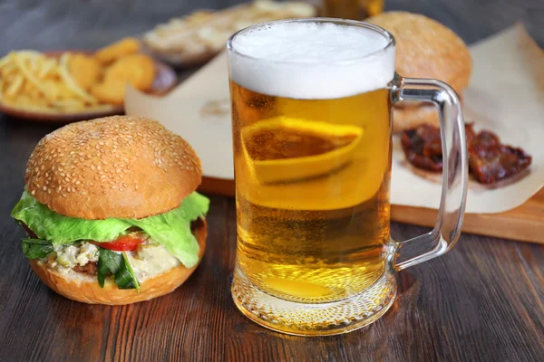 Taza de vidrio de cerveza ligera con hamburguesas en mesa de madera oscura, de cerca — Foto de Stock