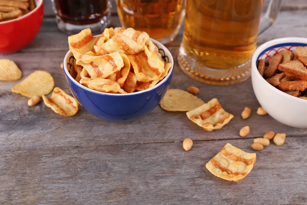 Tazas de cerveza y tazones con aperitivos en mesa de madera — Foto de Stock