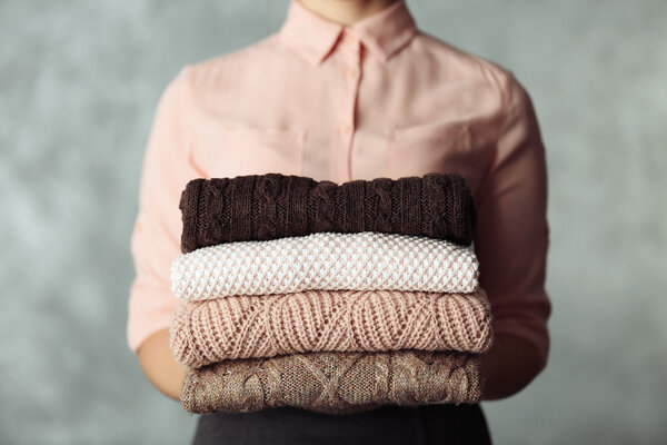 Stack of knitted clothes in female hands on grey blurred background