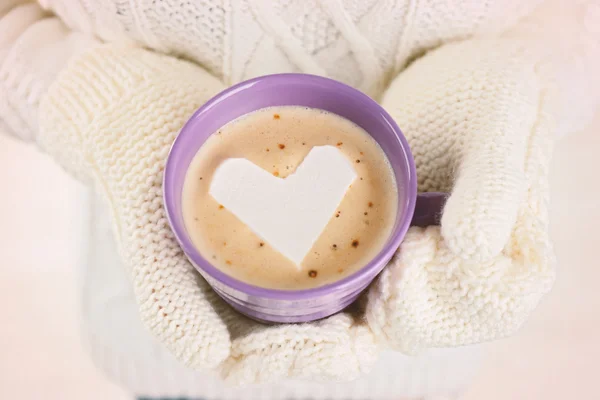 Frauenhände in warmen Fäustlingen halten Tasse heißen Cappuccino mit Herz Marshmallow, Nahaufnahme — Stockfoto