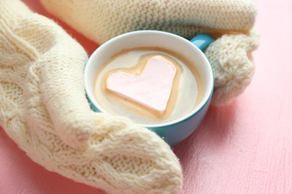 Mains féminines dans des mitaines chaudes tenant une tasse de cappuccino chaud avec guimauve cardiaque sur fond rose, gros plan — Photo