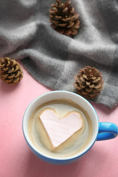 Cup of hot cappuccino with heart marshmallow and warm scarf on pink background, close up