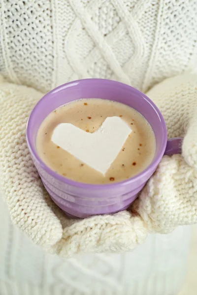 Female hands in warm mittens holding cup of hot cappuccino with heart marshmallow, close up — Stock Photo, Image