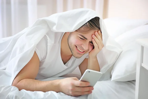 Молодой человек в постели просыпается с телефоном дома — стоковое фото