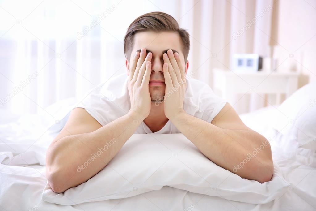 Young man closes eyes with hands in bed at home