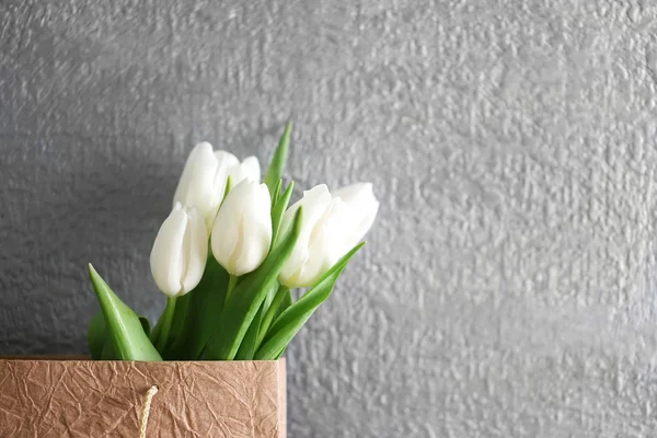 Tulipes blanches dans un sac en papier sur une table en bois contre le mur gris, gros plan — Photo