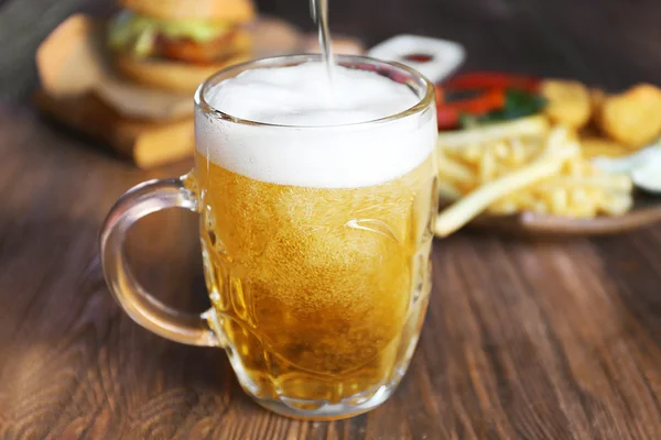 Glass mug of draft light beer with snacks on dark wooden table, close up — Stock Photo, Image