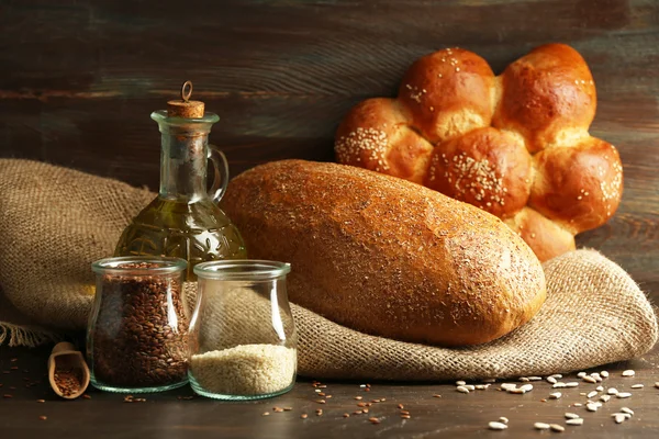 Composition of seeds in banks, bread, buns and oil on wooden table background, closeup — Stock Photo, Image