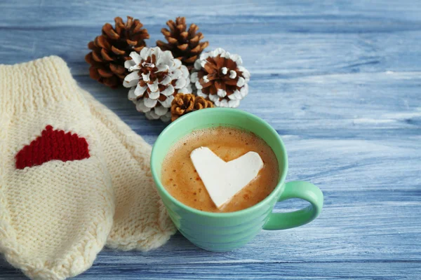 Xícara de cappuccino quente com marshmallow coração, mitenes quentes e cones de pinho em fundo de madeira azul, close up — Fotografia de Stock