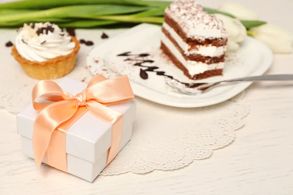 Sweet creamy cake with flowers and present box on wooden table closeup — Stock Photo, Image