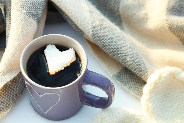 Cup of hot coffee with heart marshmallow and warm clothes on snowy background, close up — Stock Photo, Image