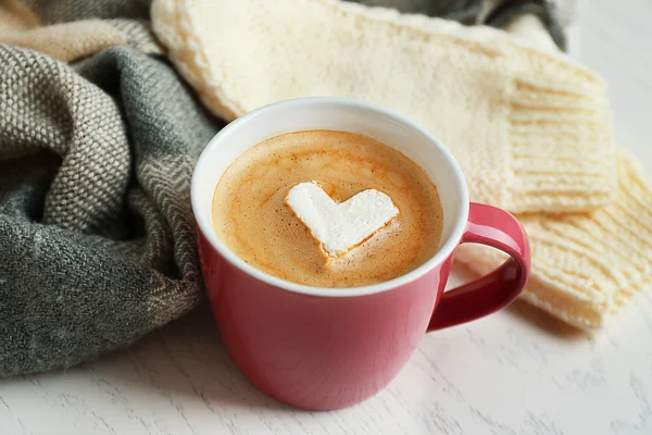 Tasse heißen Cappuccino mit Herz-Marshmallow und warme Kleidung auf hellem Holzhintergrund, Nahaufnahme — Stockfoto