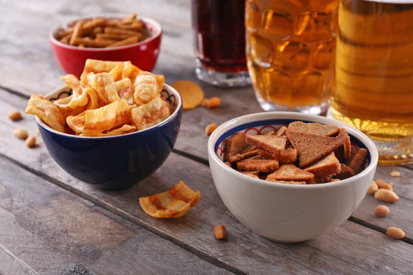 Mugs of beer and bowls with snacks on wooden table — Stock Photo, Image