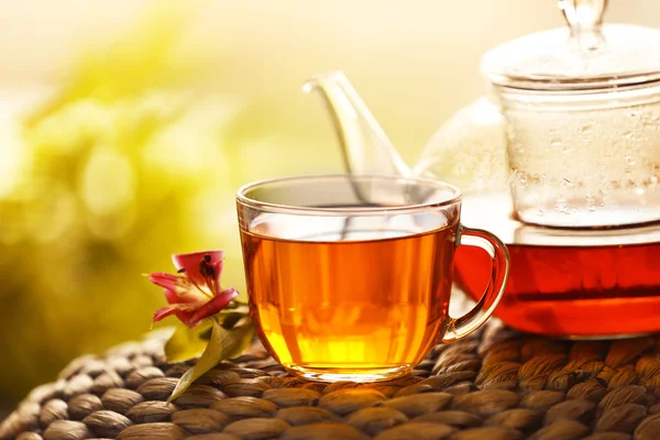 Glass cup of tea with teapot on wicker mat against blurred background — Stock Photo, Image