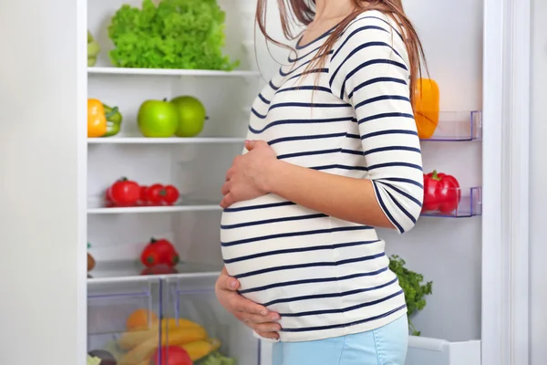 妊娠中の女性が台所でオープン冷蔵庫の近くに立って — ストック写真