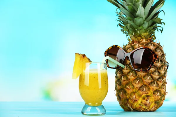 Ripe juicy pineapple in sun glasses drinking smoothie from glass on blue blurred background — Stock Photo, Image