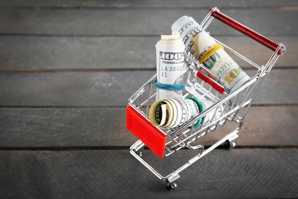 Pequeño carrito de compras con billetes en dólares sobre fondo de madera — Foto de Stock