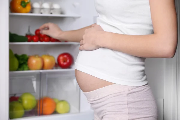 Gravid kvinna som står nära kylskåp fulla av hälsosam mat — Stockfoto