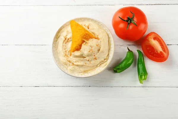 Ceramic bowl of tasty hummus with chips, chili and tomato on table — Stock Photo, Image