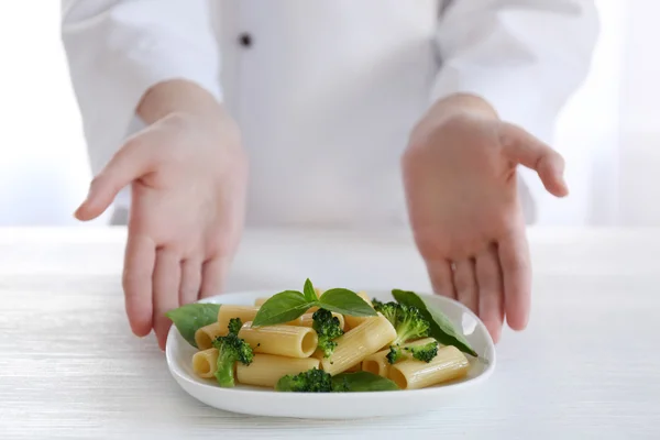 Köchin hält Teller mit gekochten Rigatoni-Nudeln mit Brokkoli und Basilikum am Tisch — Stockfoto