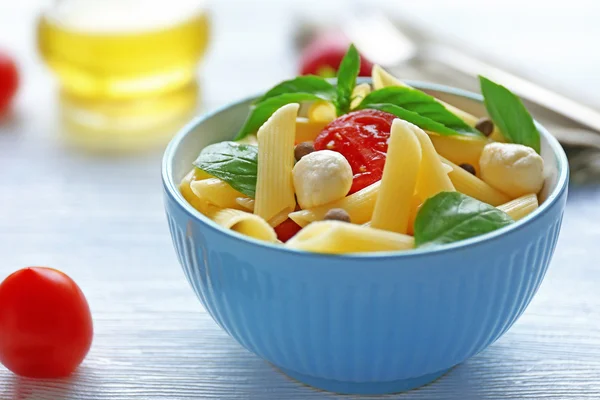 Cooked penne pasta with mozzarella, fresh tomatoes and basil in blue bowl on wooden table — Stock Photo, Image