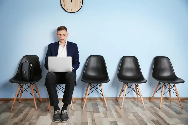 Young man in suit sitting on chair with laptop and waiting for job interview against blue wall background