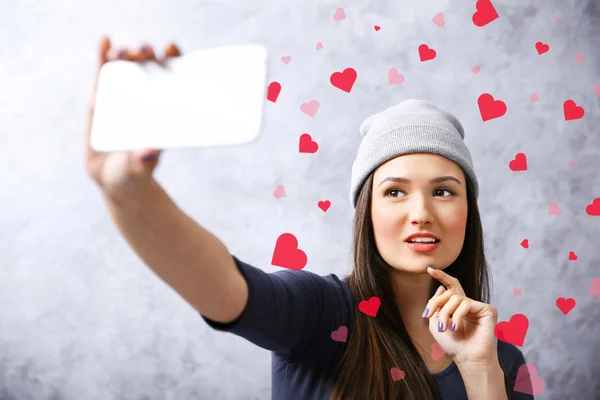Young girl in hat taking photo of her self with smart phone on grey wall background — Stock Photo, Image