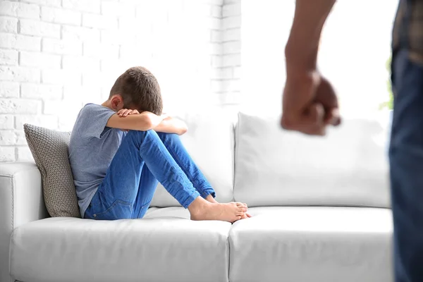 Family problems concept. Frightened boy sitting on sofa and father with bunched fist