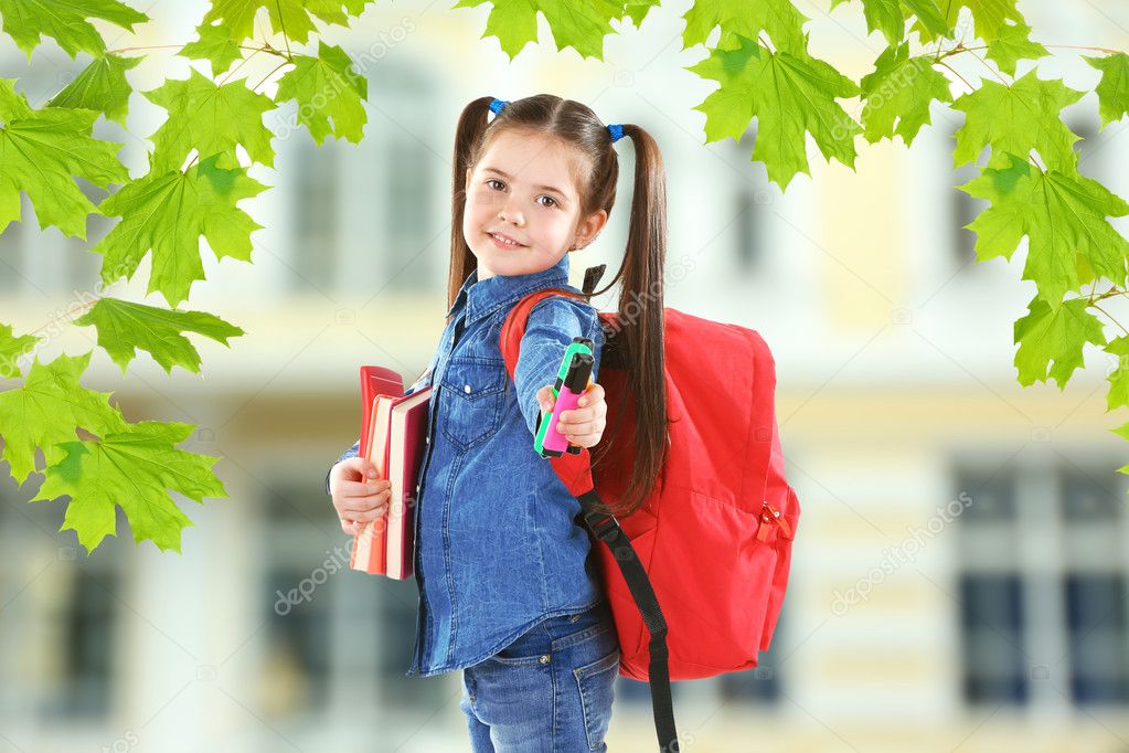 Little girl with red back pack holding books and stationery on blurred school building background
