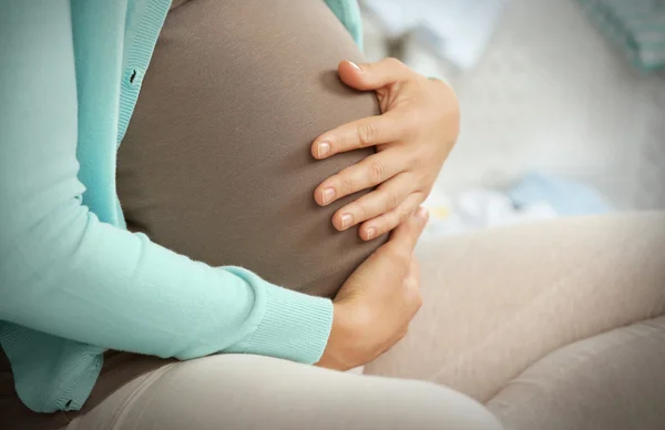 Kobieta w ciąży, trzymając ręce na brzuchu i siedzi na kanapie w pokoju — Zdjęcie stockowe