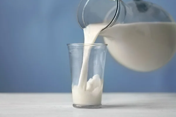 Mléko se vylévá ze džbánu do sklenice na modrém pozadí — Stock fotografie