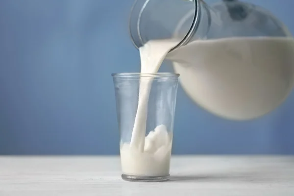 Leche vertida de una jarra en un vaso sobre fondo azul —  Fotos de Stock