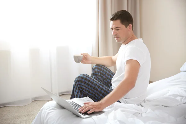 Bonito homem sentado na cama com laptop e xícara de café — Fotografia de Stock