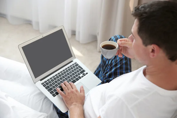 Hombre guapo sentado en la cama con portátil y taza de café —  Fotos de Stock