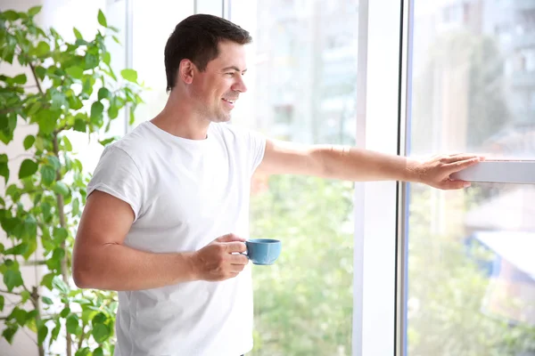 Beau homme debout près de la fenêtre avec une tasse de café du matin — Photo