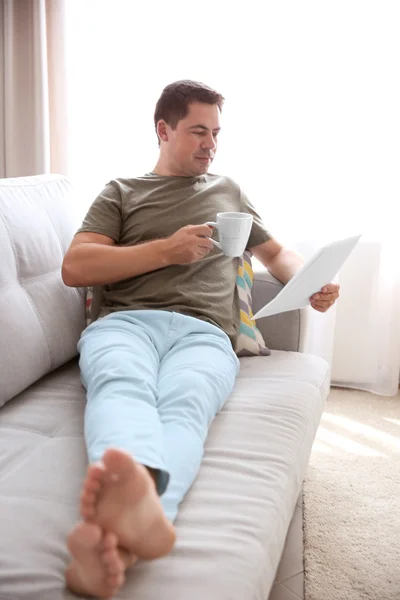 Hombre guapo leyendo en el sofá con una taza de café —  Fotos de Stock