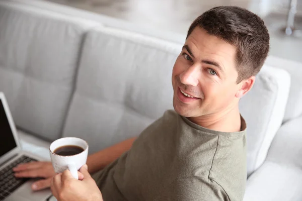 Hombre guapo sentado en el sofá con portátil y taza de café —  Fotos de Stock