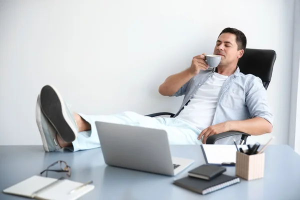 Relajado guapo hombre está bebiendo café después de haber puesto sus piernas en la mesa — Foto de Stock