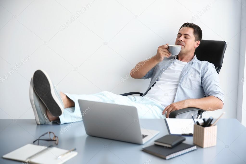 Relaxed handsome man is drinking coffee having put his legs on table