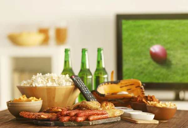 Snack gustosi e bottiglie con birra sul tavolo della cucina sullo sfondo della TV offuscata — Foto Stock