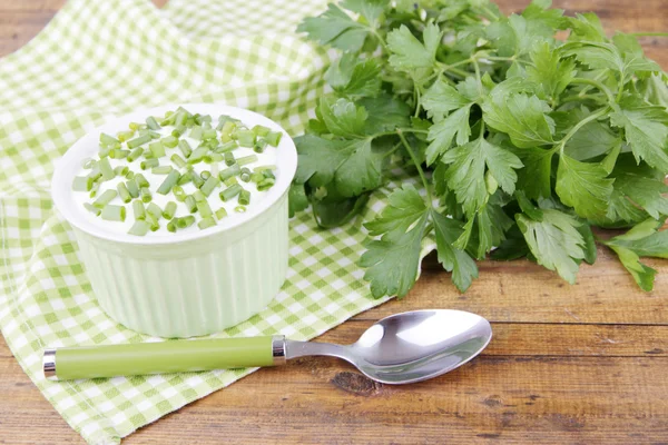 Plastic round bowl of cream with a tuft of parsley near it on a napkin on wooden background — Stock Photo, Image