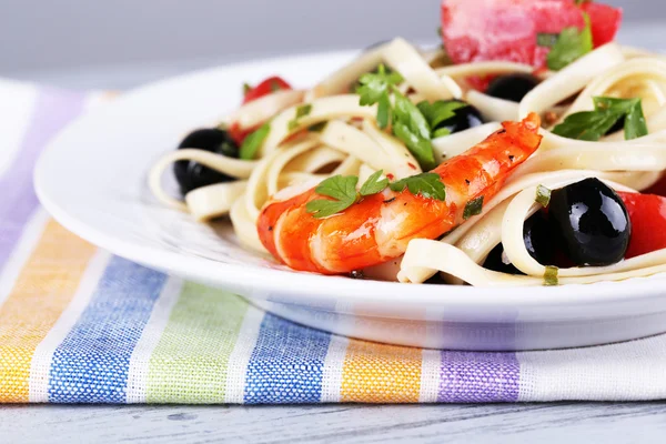 Gamberi freschi con spaghetti, olive, pomodori e prezzemolo in un grande piatto rotondo su un tovagliolo su un tavolo di legno su sfondo grigio — Foto Stock