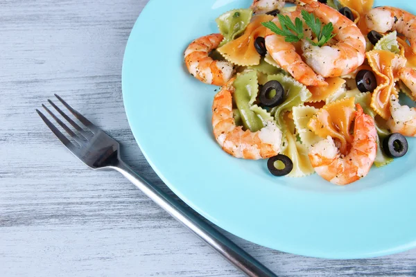 Fresh prawns with coloured macaroni, olives and parsley in a big blue round plate with a fork on wooden background — Stock Photo, Image